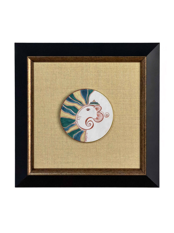 Framed circular ceramic medallion hand-painted with a stylized Ganesha profile in teal, gold and terracotta rays, mounted on golden linen in a black and gold frame — Tangled Monk original hand-drawn art